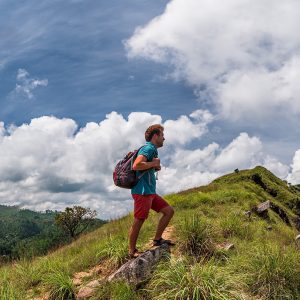 Caucasian hiker man take a rest on mountain peak with backpack. He is looking on beautiful landscape with blue sky and green hills. Panorama view