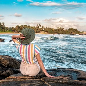 Young woman in knitting hat, sitting on the ocean rocky shore, and looks at the waves. Vacation theme